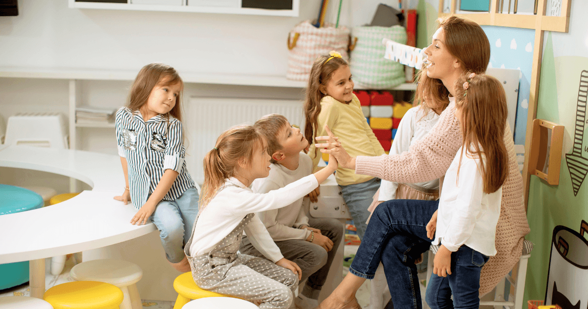 A group of young children smile and interact with a woman, likely a teacher, who is sitting and high-fiving one of them in a colorful classroom, embodying trauma-informed teaching practices.