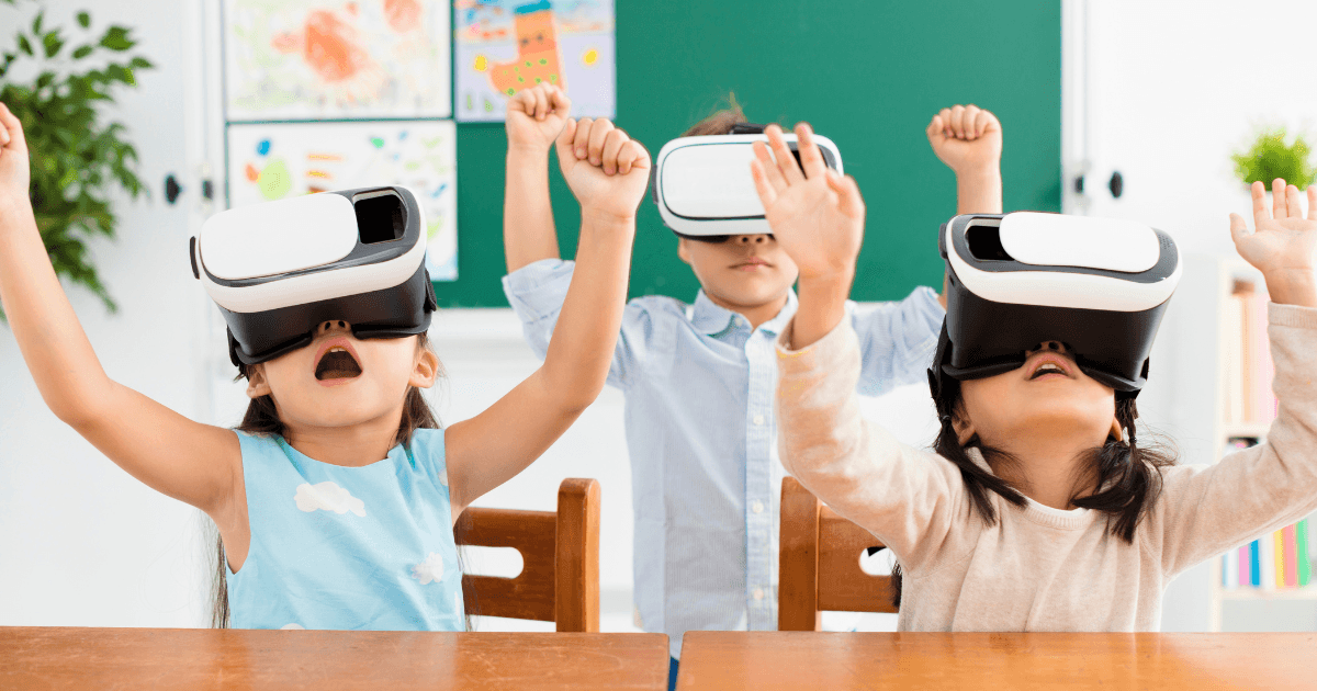 Three children wearing virtual reality headsets sit at desks in a classroom with their hands raised excitedly, appearing engaged and amazed by the virtual field trips they're experiencing.