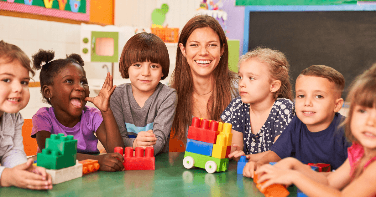 A smiling teacher sits at a table with five young children playing with colorful plastic building blocks in a classroom setting. The kids appear happy and engaged in the activity, which can be observed through in-person visits or virtual tours.