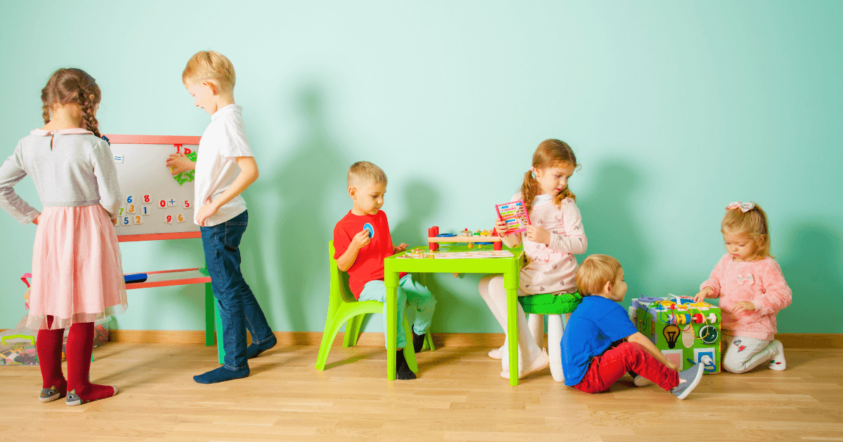 Six young children play and learn in a brightly lit room, exploring activities perfect for what age for pre-K vs preschool. Some use educational toys at a table, others work with numbers on a board. The background wall is light blue.