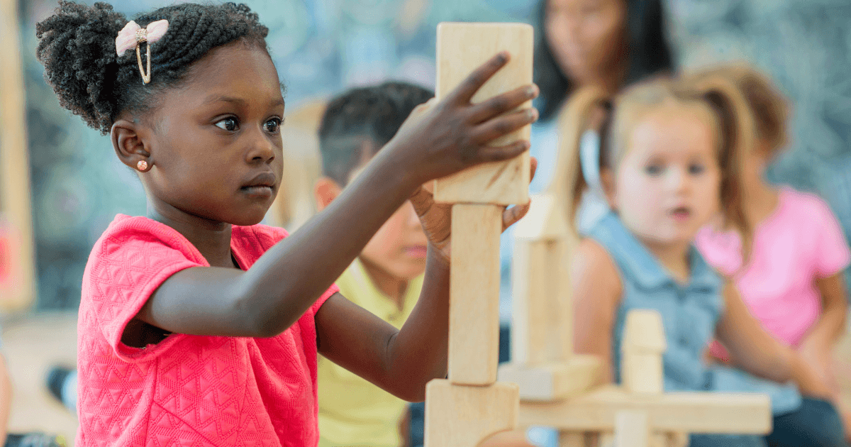 A young girl wearing a pink shirt carefully stacks wooden blocks in a classroom inspired by the Reggio Emilia approach, with other children blurred in the background, watching or participating in similar activities.