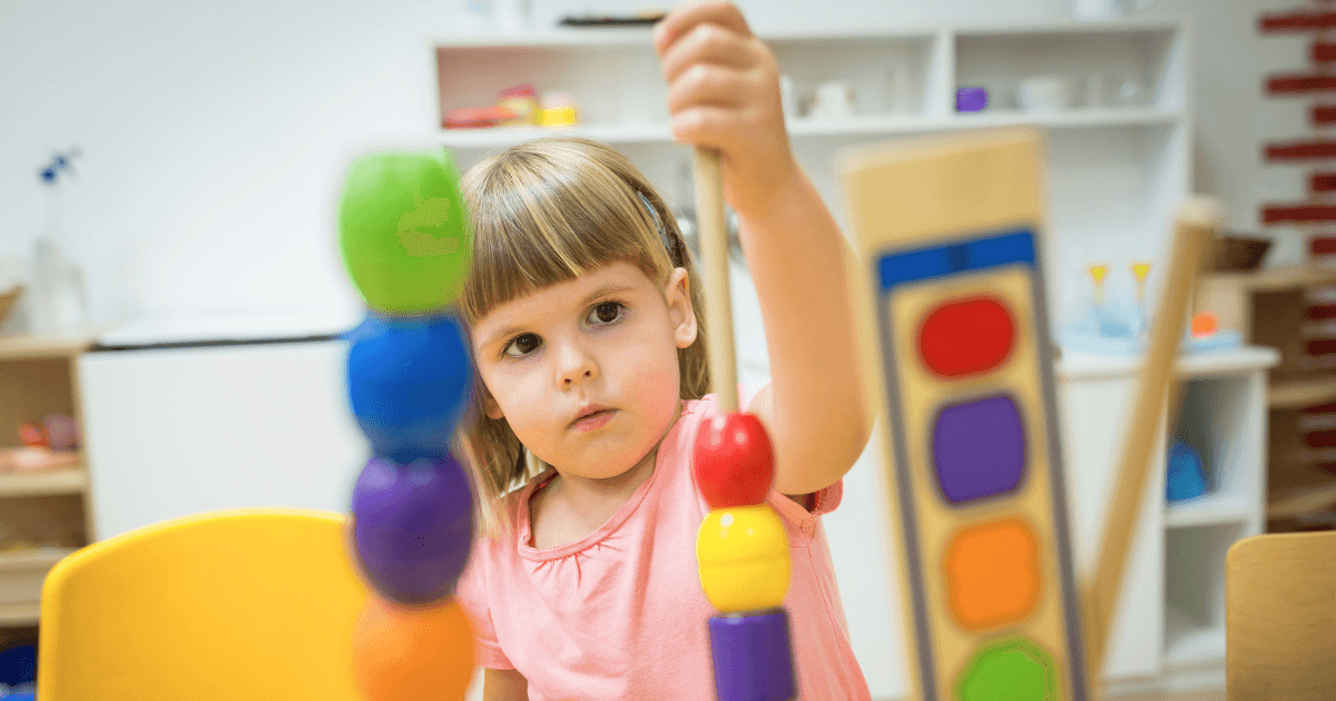 A young girl in a pink shirt stacks colorful wooden beads on a rod in a classroom, focusing intently—an example of what is play-based learning, where children develop skills through engaging, hands-on activities.