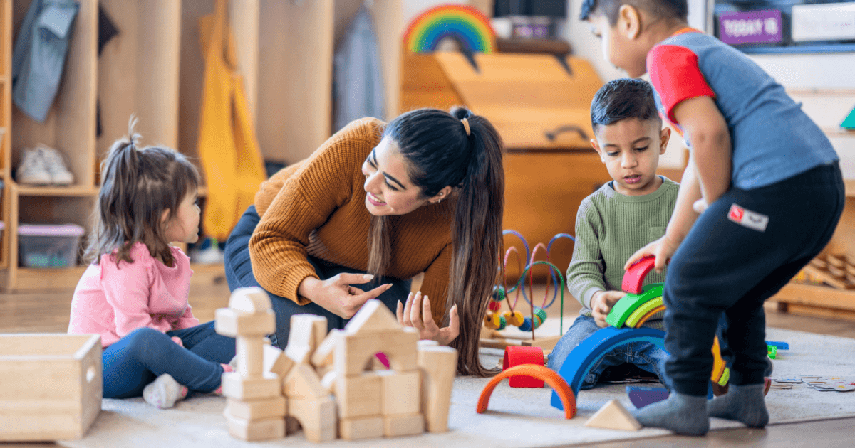 A woman sits on the floor smiling and talking to three young children, playing with wooden blocks and colorful toys in a bright, cheerful room—an engaging setting for exploring what should my child know before kindergarten.