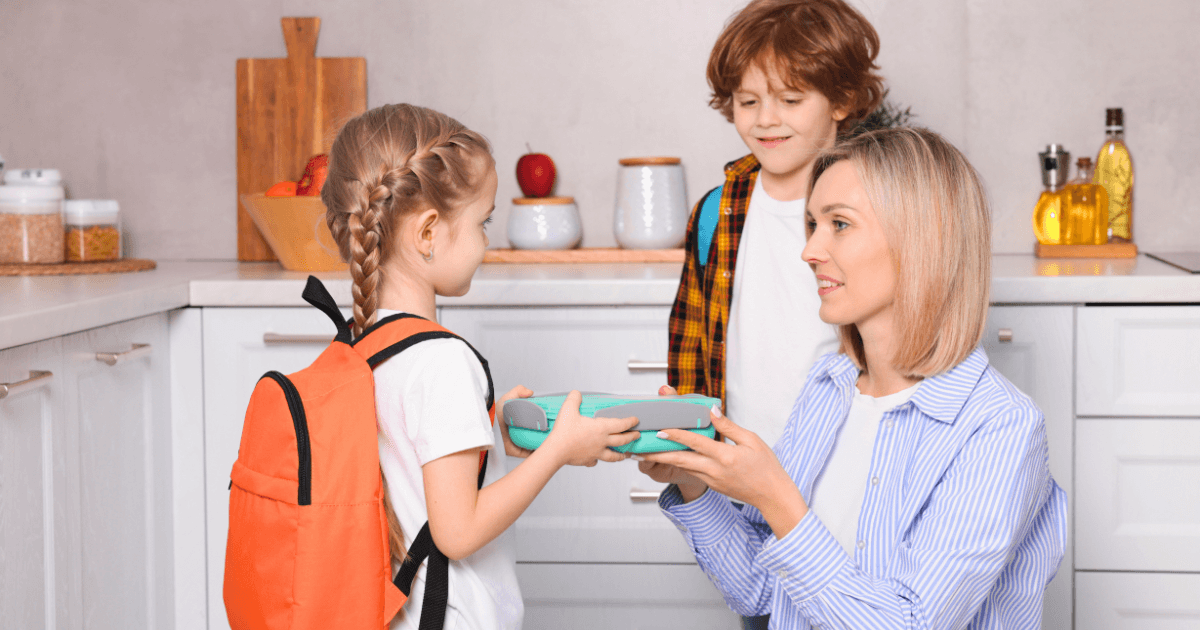 A woman kneels in a kitchen, handing a lunchbox to a young girl wearing an orange backpack, while a young boy stands nearby—both children ready for school as she wonders, "When should my child start preschool?.