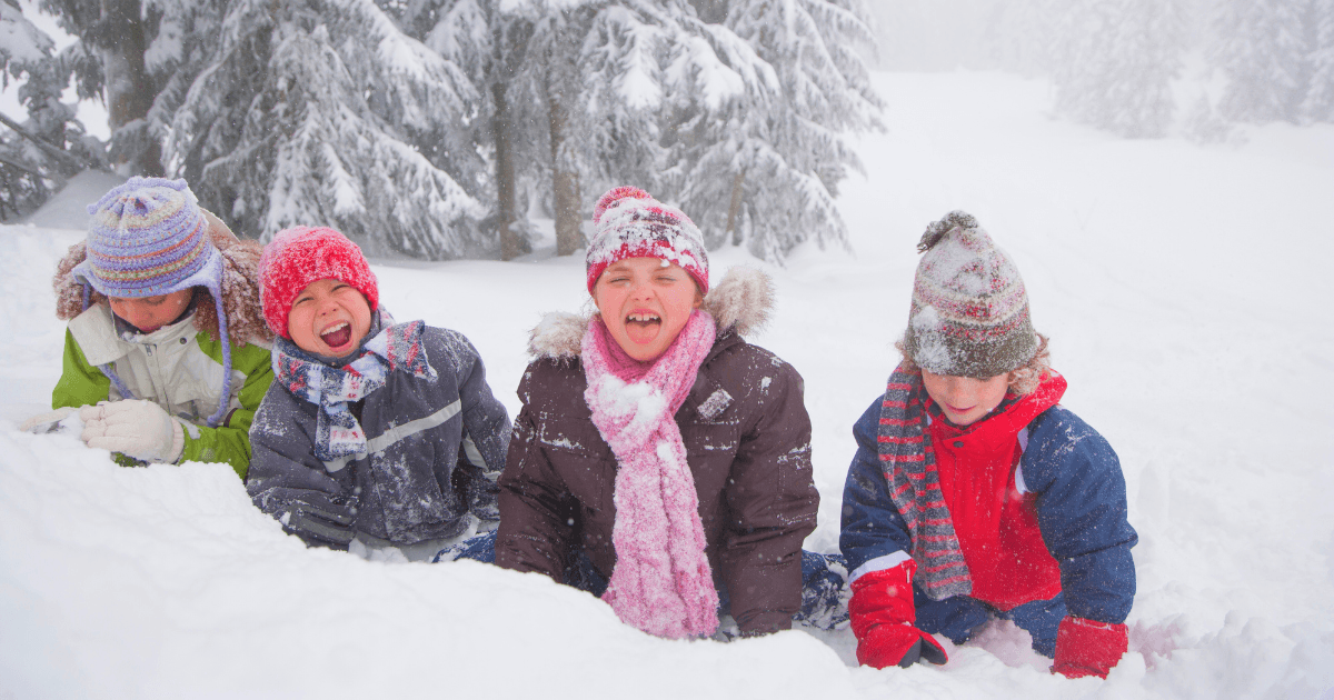 Four children bundled in winter coats, scarves, and hats sit together in the snow, smiling and laughing after fun winter STEM activities. Snow-covered trees and a white, wintry landscape are in the background.
