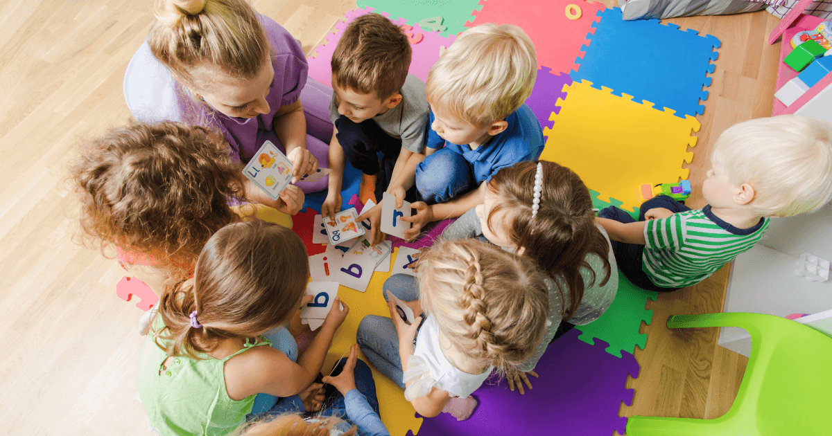 A group of young children sit in a circle with a teacher on colorful foam mats, playing with alphabet cards and enjoying a year of preschool activities as they learn letters together in a bright classroom.