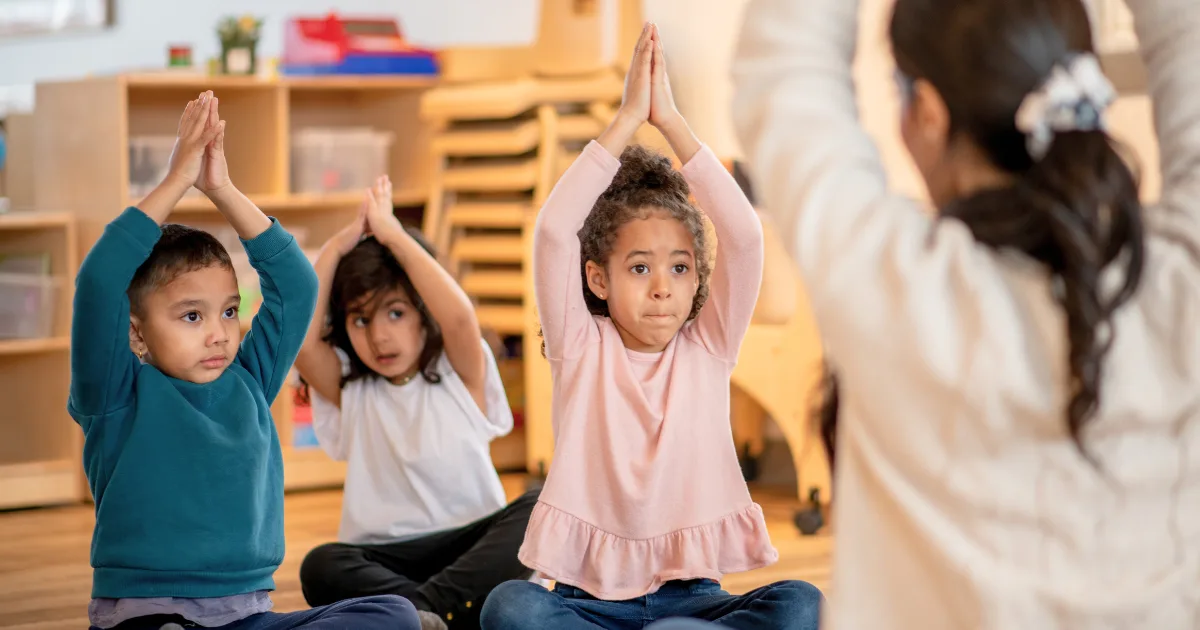 Three young children sit cross-legged on the floor with their arms raised above their heads, following a woman’s lead in a yoga session. Shelves and stacked chairs are visible in the classroom background.